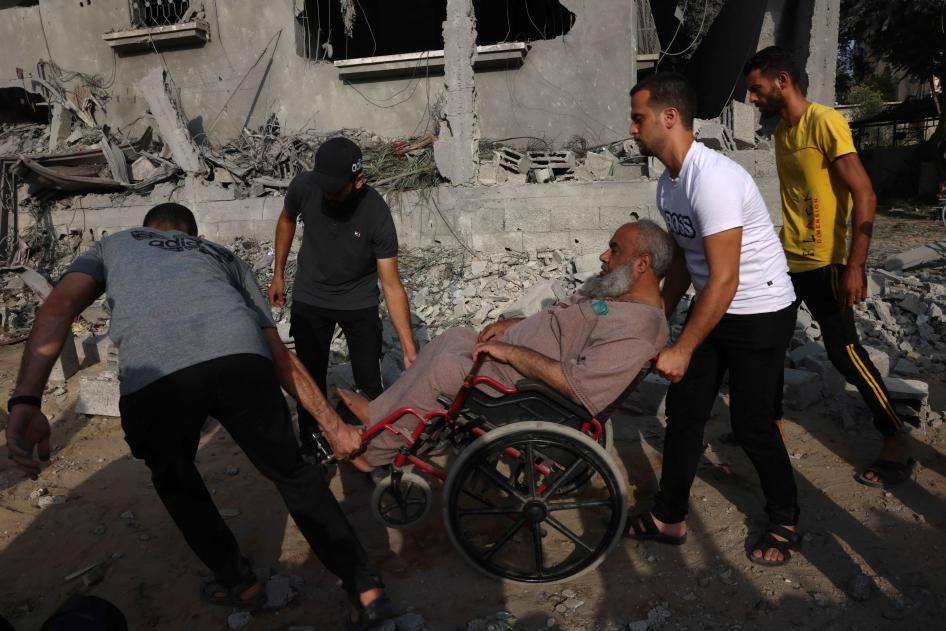 Tiga orang pria membantu seorang pria berkursi roda bergerak di tengah reruntuhan dan kehancuran bangunan di Jalur Gaza selatan pada 22 Oktober 2023. 