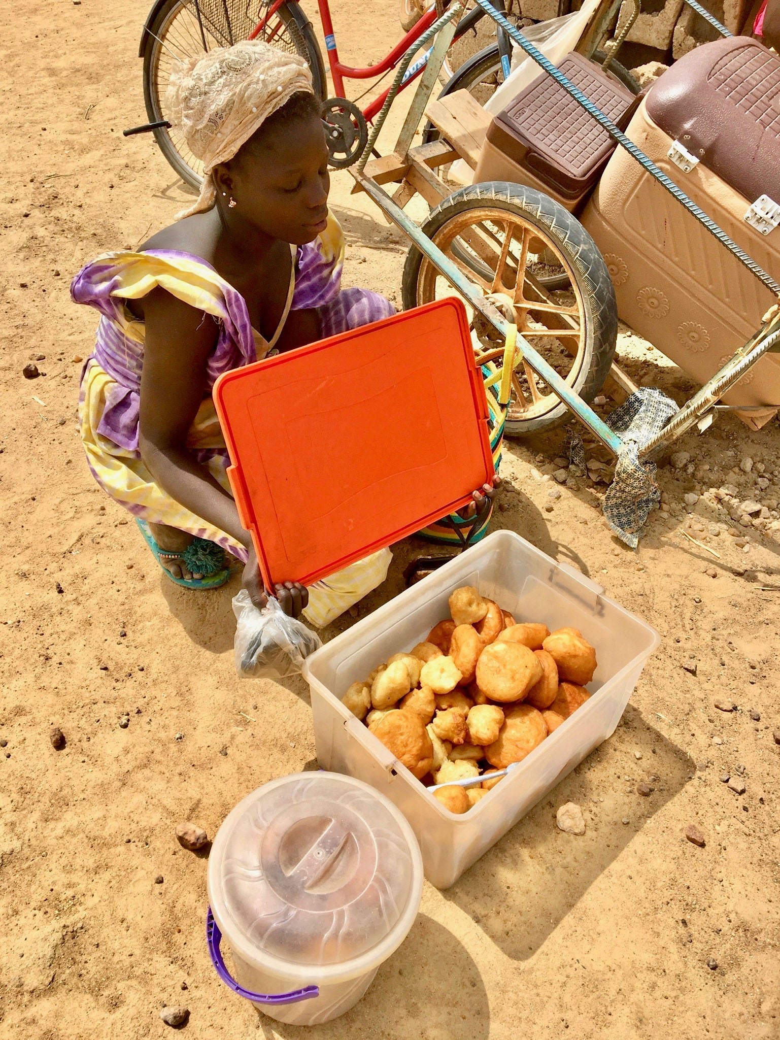 A girl kneels on the ground while displaying a box of cakes for sale