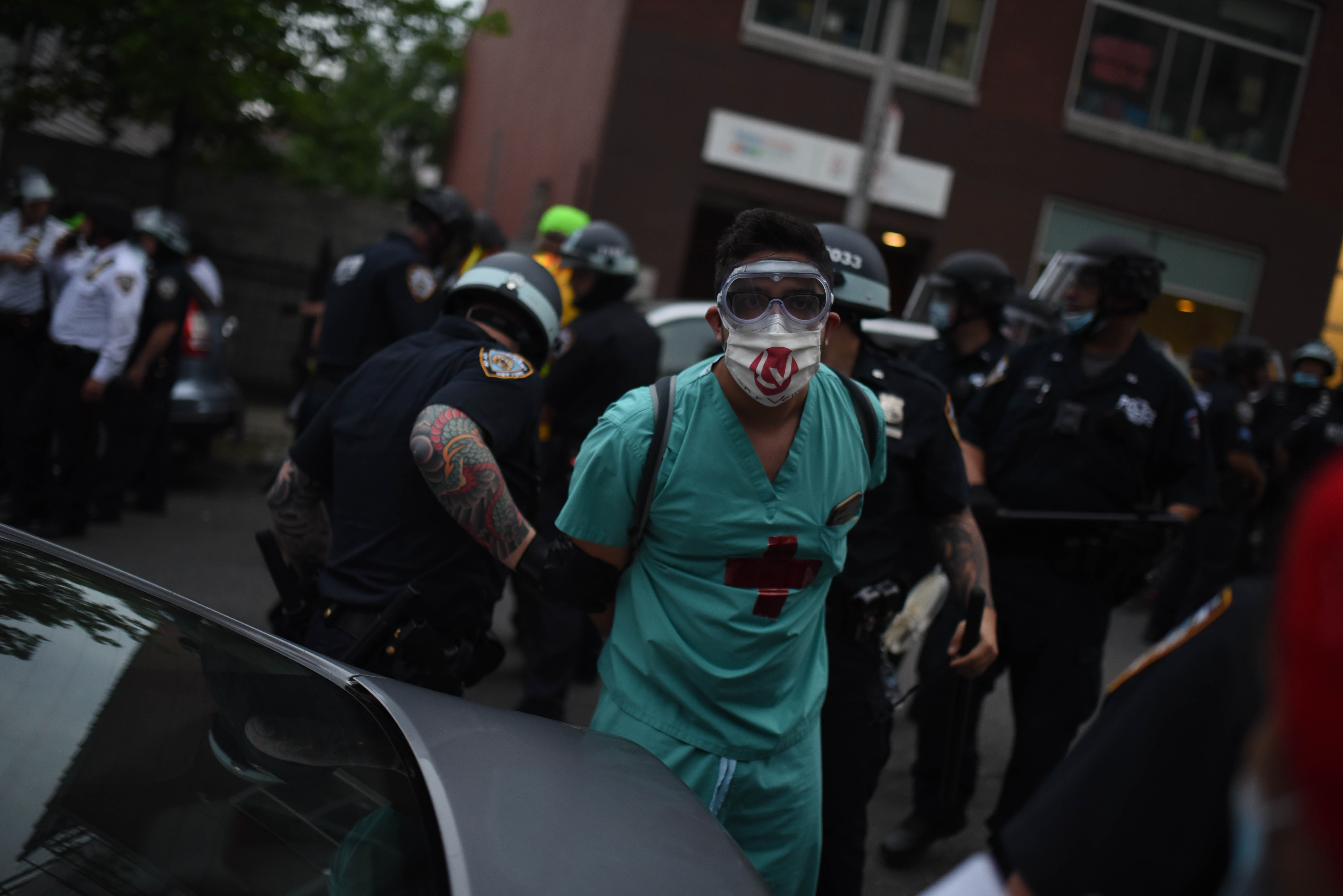 A man in a mask and hospital scrubs is handcuffed by police officers