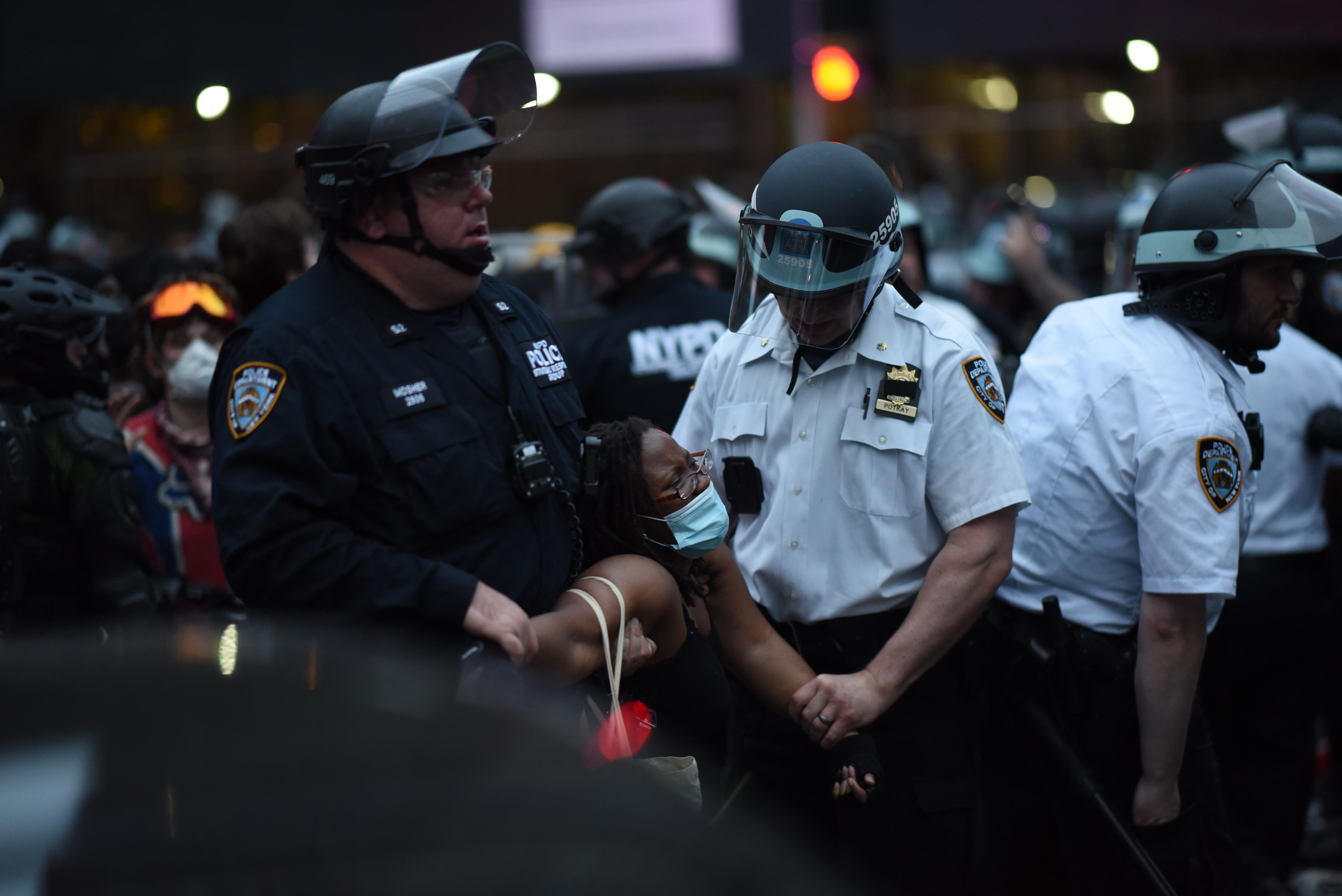 A Black woman in a mask is forcibly detained by two police officers