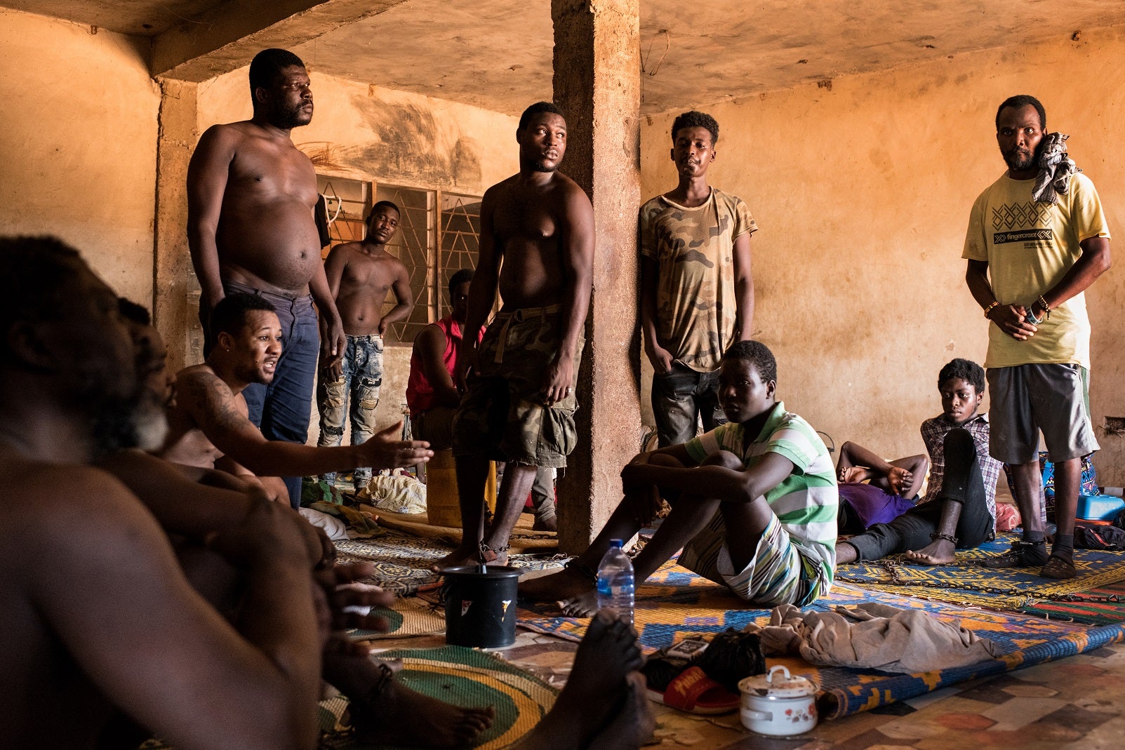 A group of men sitting and standing in a crowded room, some with chains around their ankles.