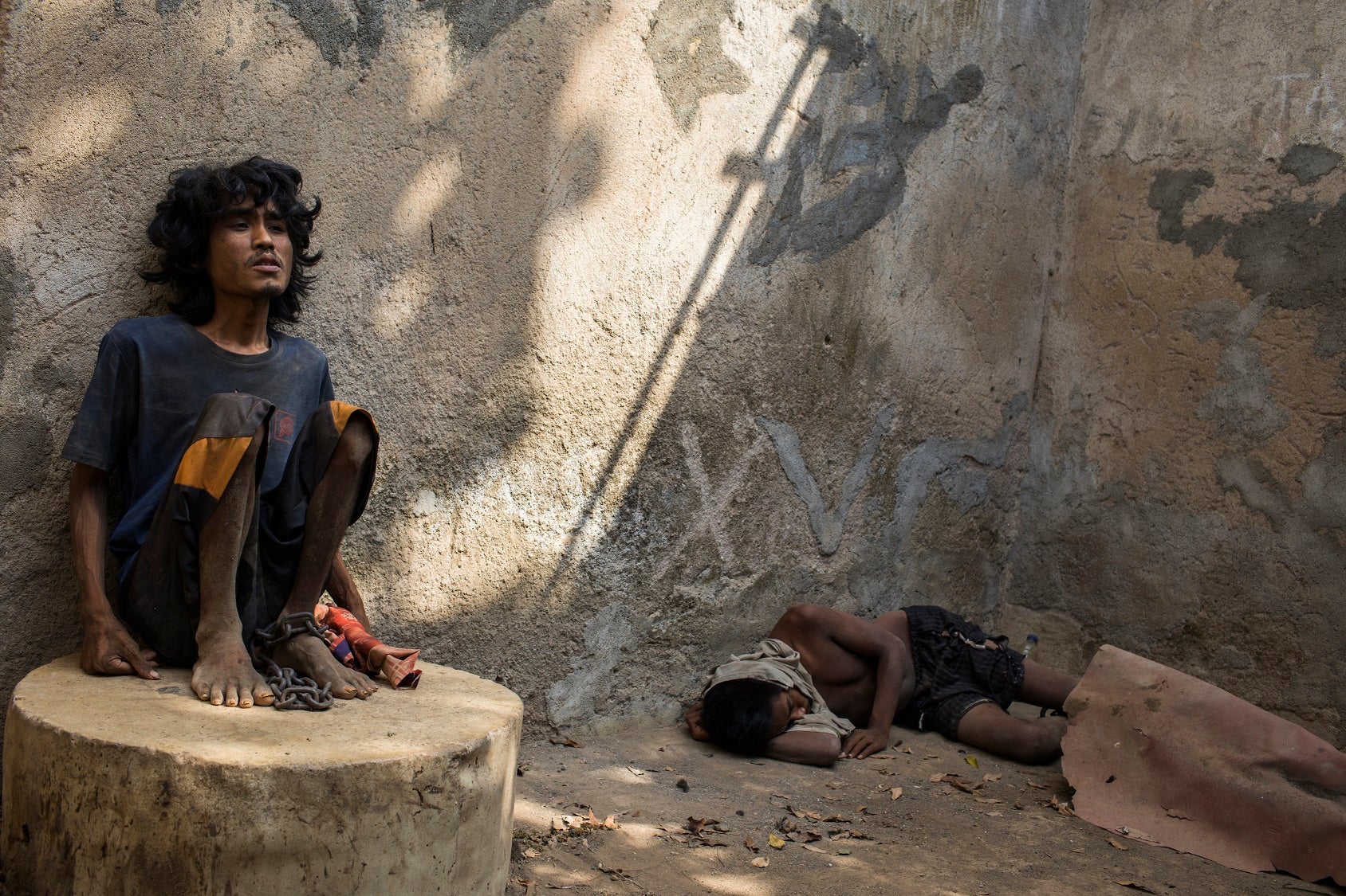 A man sitting on a concrete structure with a chain around his ankle. Another man is lying on the concrete floor.