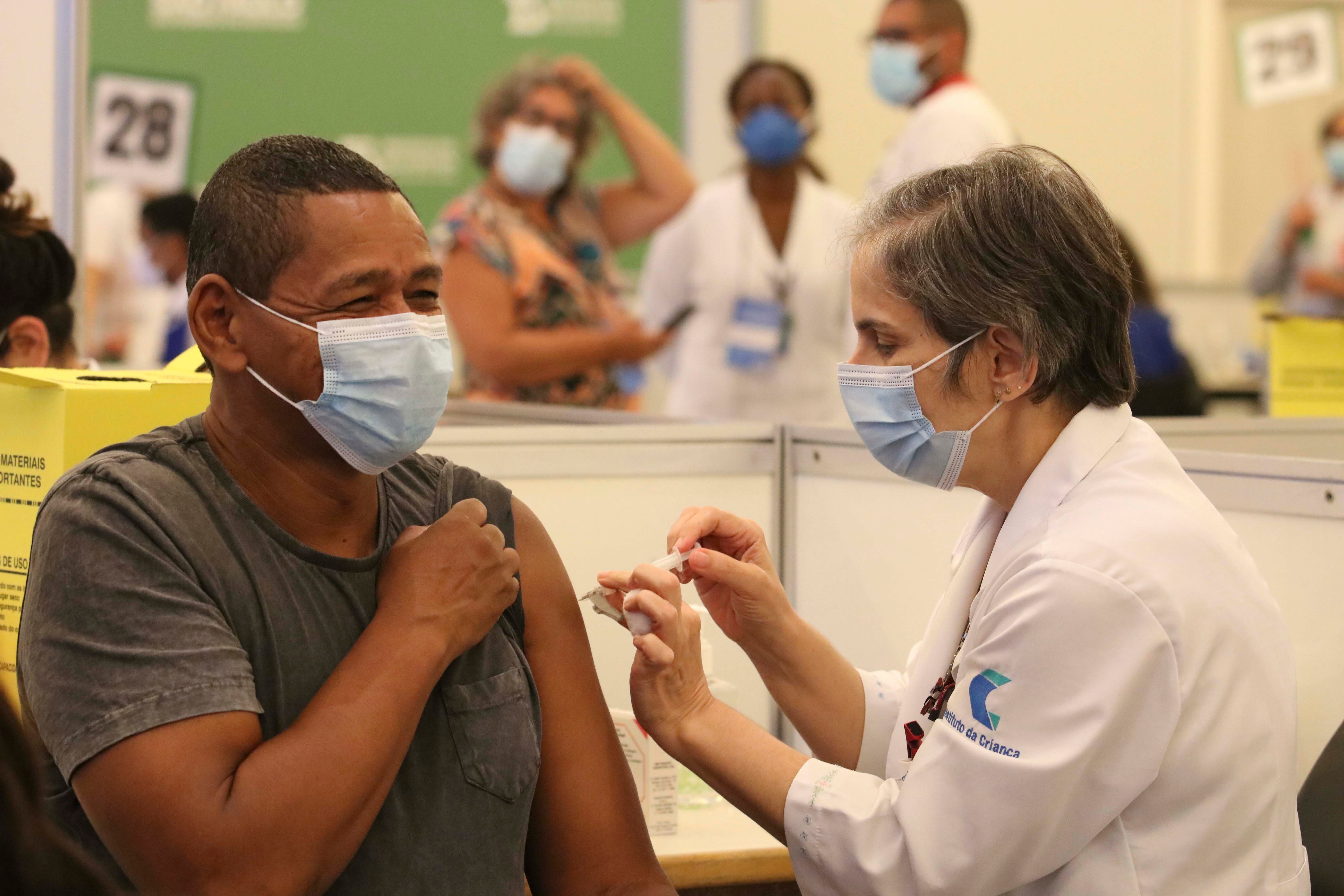 Uma mulher profissional da saude, branca, usando mascara, aplicando a vacina de covid-19 em um profissional da saúde, homem, negro, usando mascara, que está sorrindo