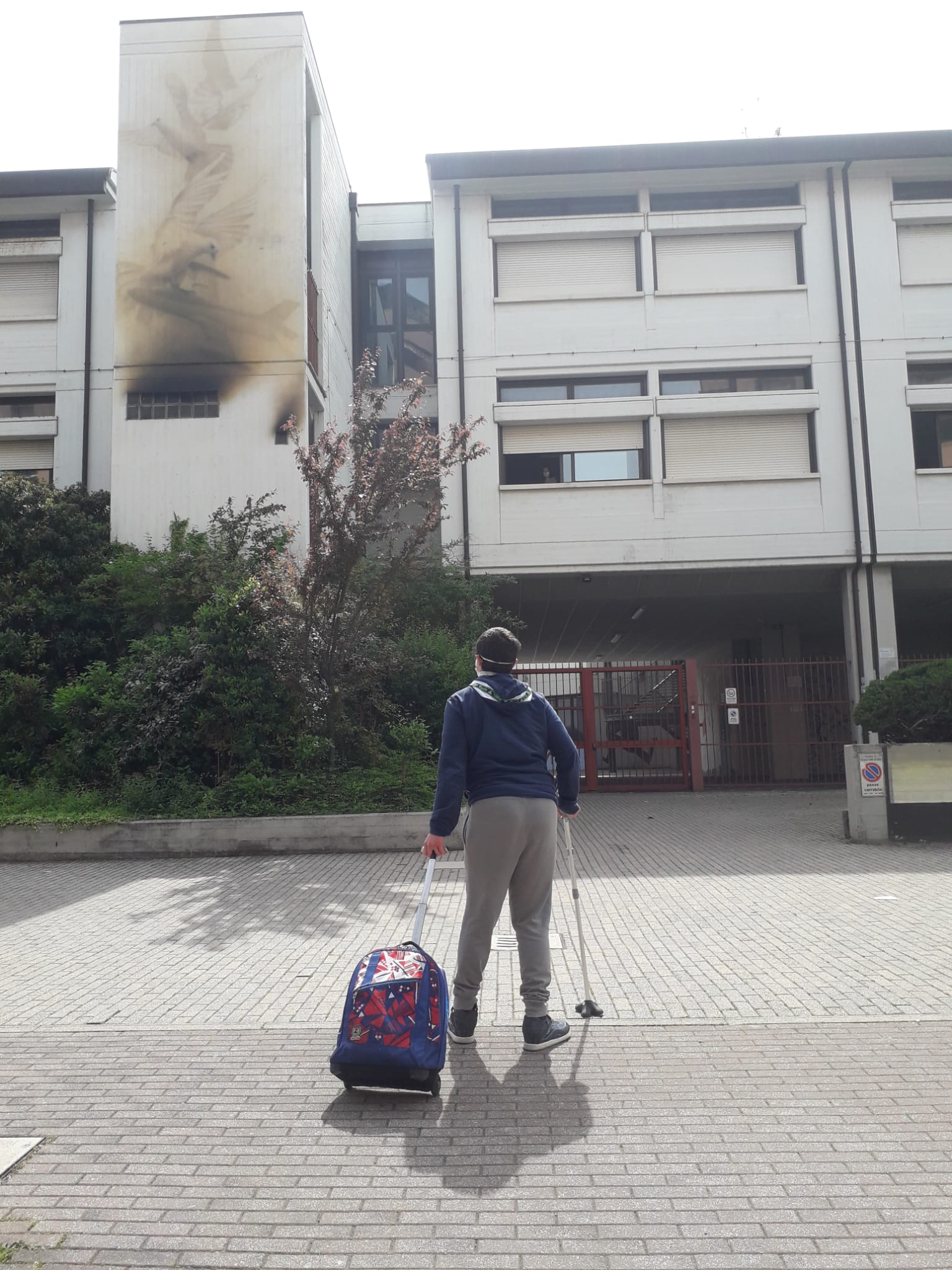 A boy carrying a backpack walks towards a building using a cane. 