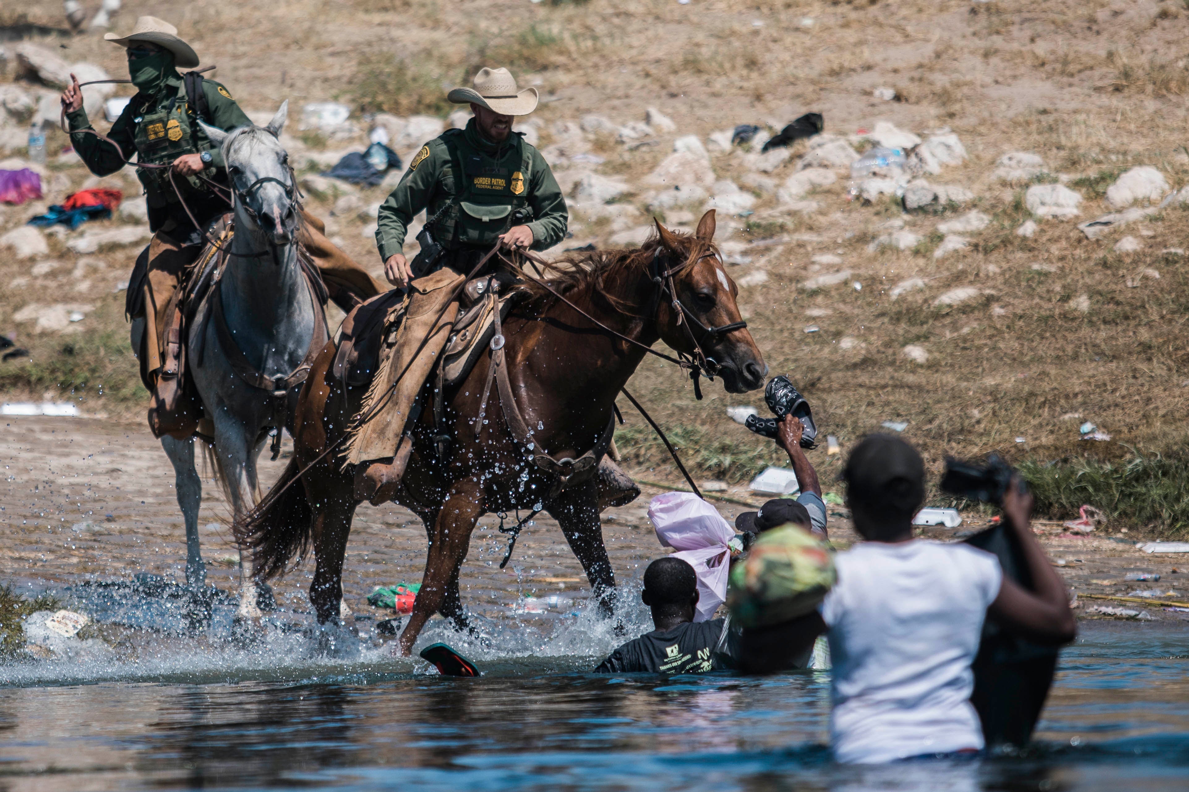 Two Customs and Border Protection agents on horseback approach three Haitian migrants standing in the river. The migrants are holding bags and shoes.