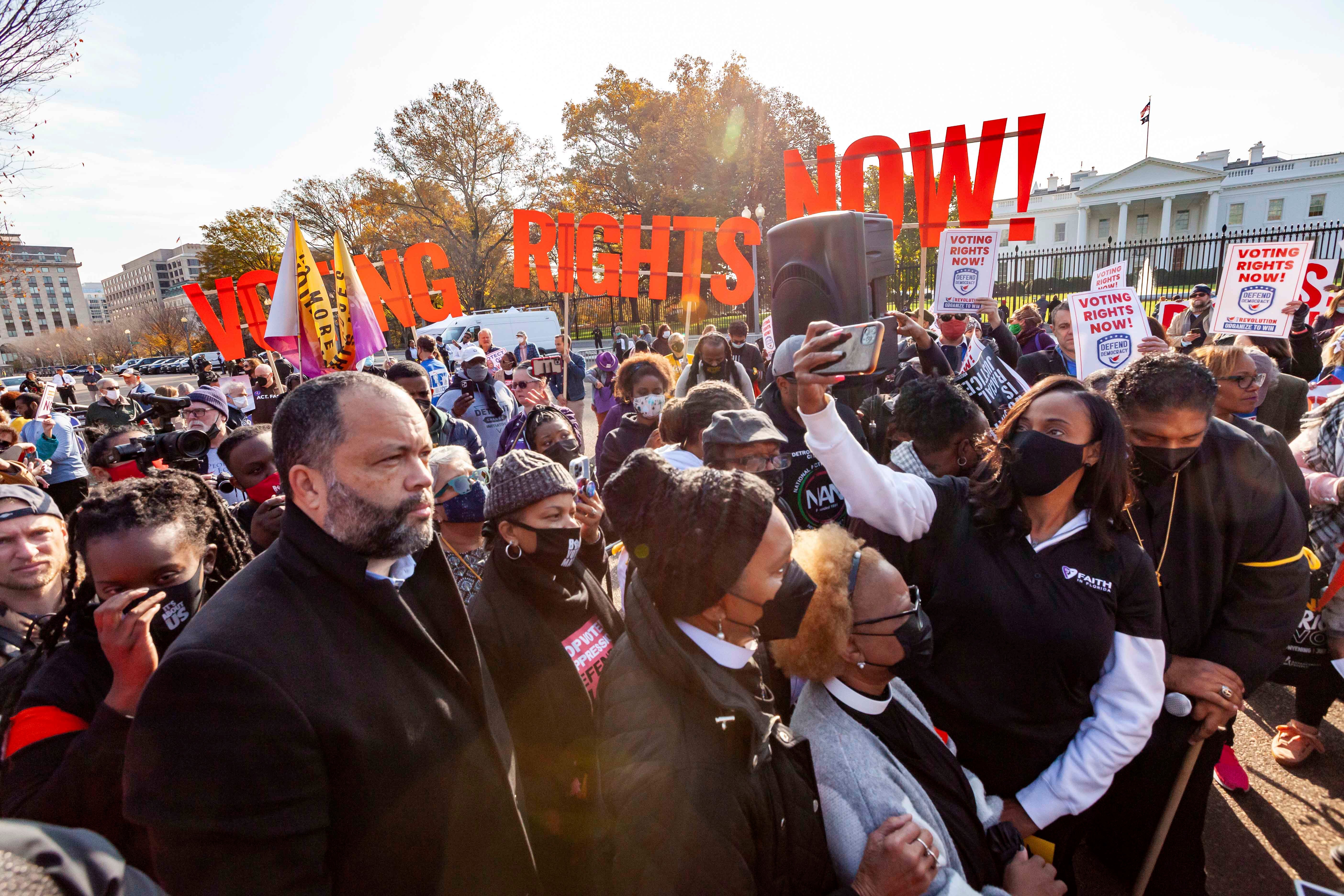 A crowd standing in front of a sign that reads "Voting Rights Now!"