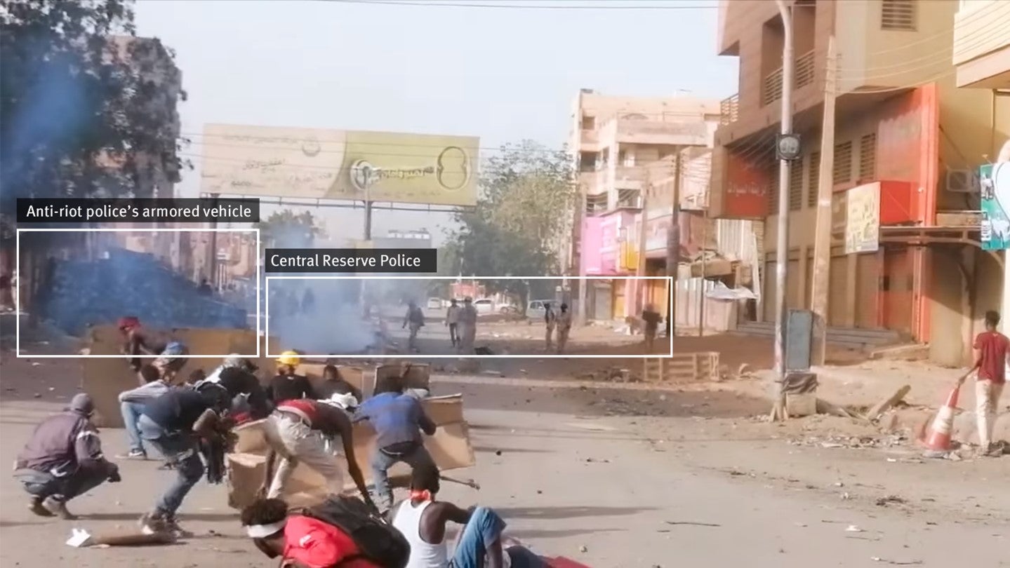 لقطة من فيديو نشر على فيسبوك. في المقدمة، يستخدم متظاهرون الورق المقوى كدرع. في الخلفية، تظهر سيارة مصفحة رباعية الدفع مع حوالي 10 عناصر من "قوات الاحتياطي المركزي" في بزات كاكية مموهة يقفون قريبا.