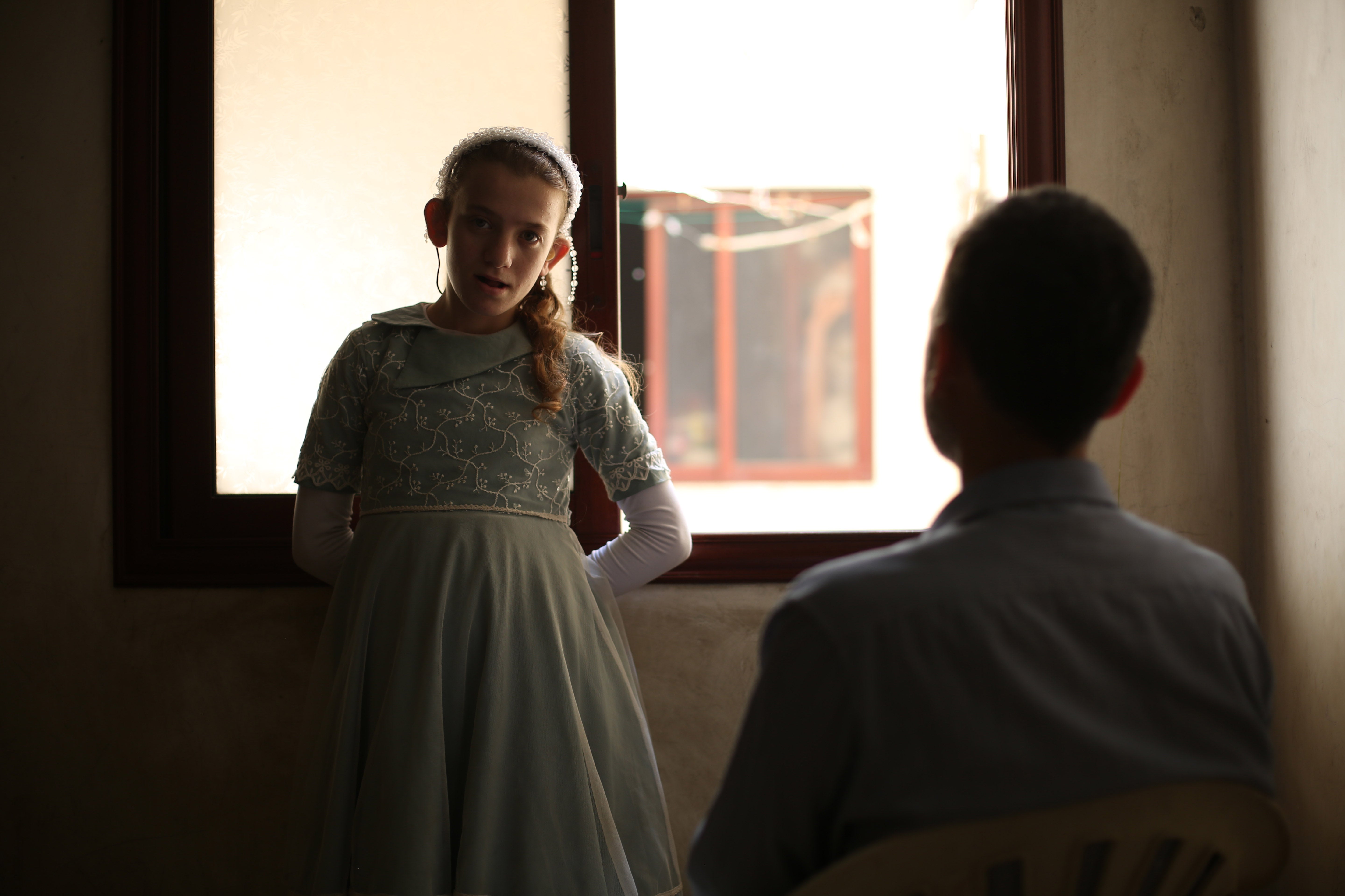 A girl stands in front of a window, looking at the camera