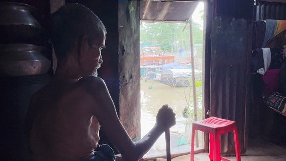 An older man sits inside his house, near a river