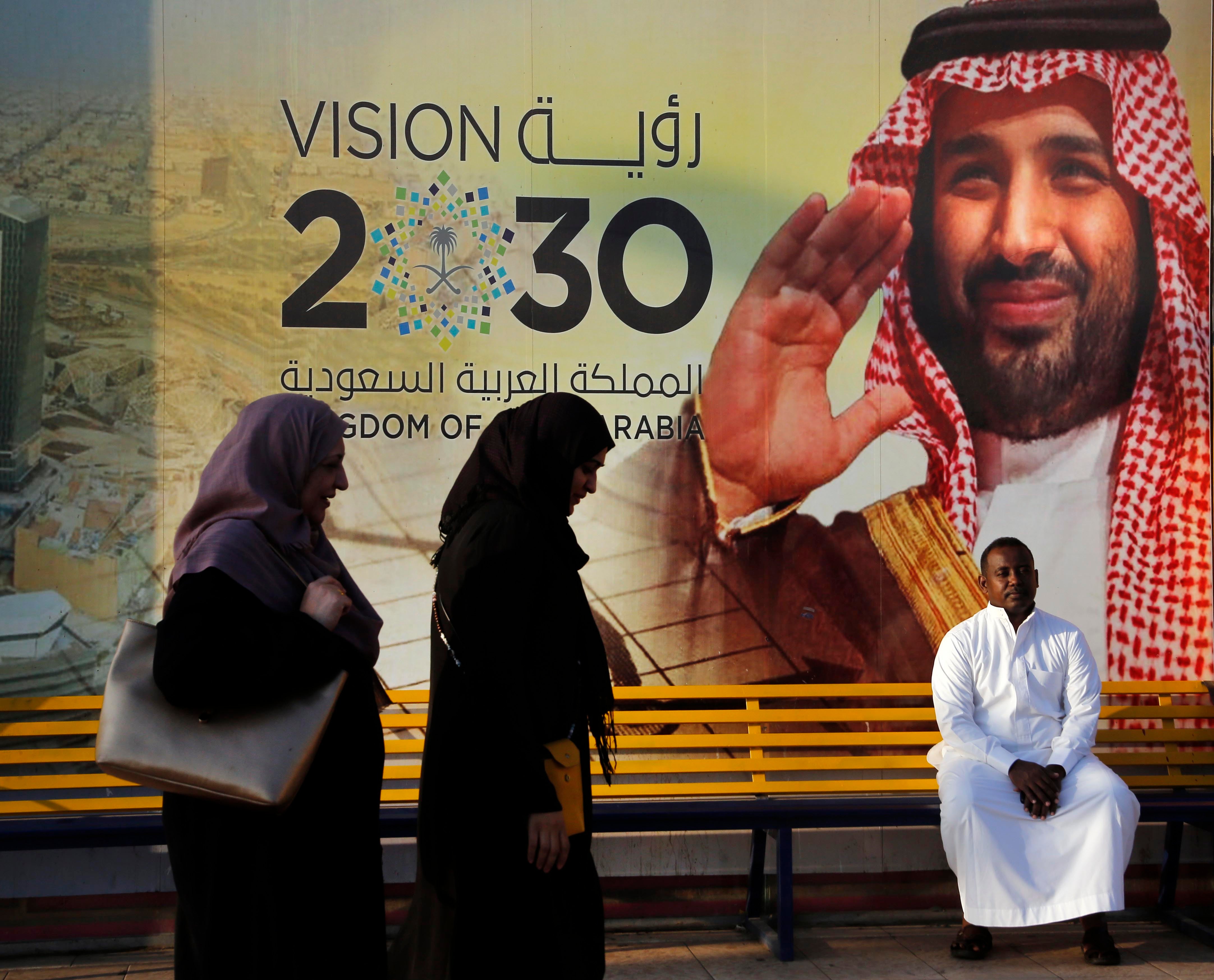 People walk past a Vision 2030 banner showing Saudi Crown Prince Mohammed bin Salman.