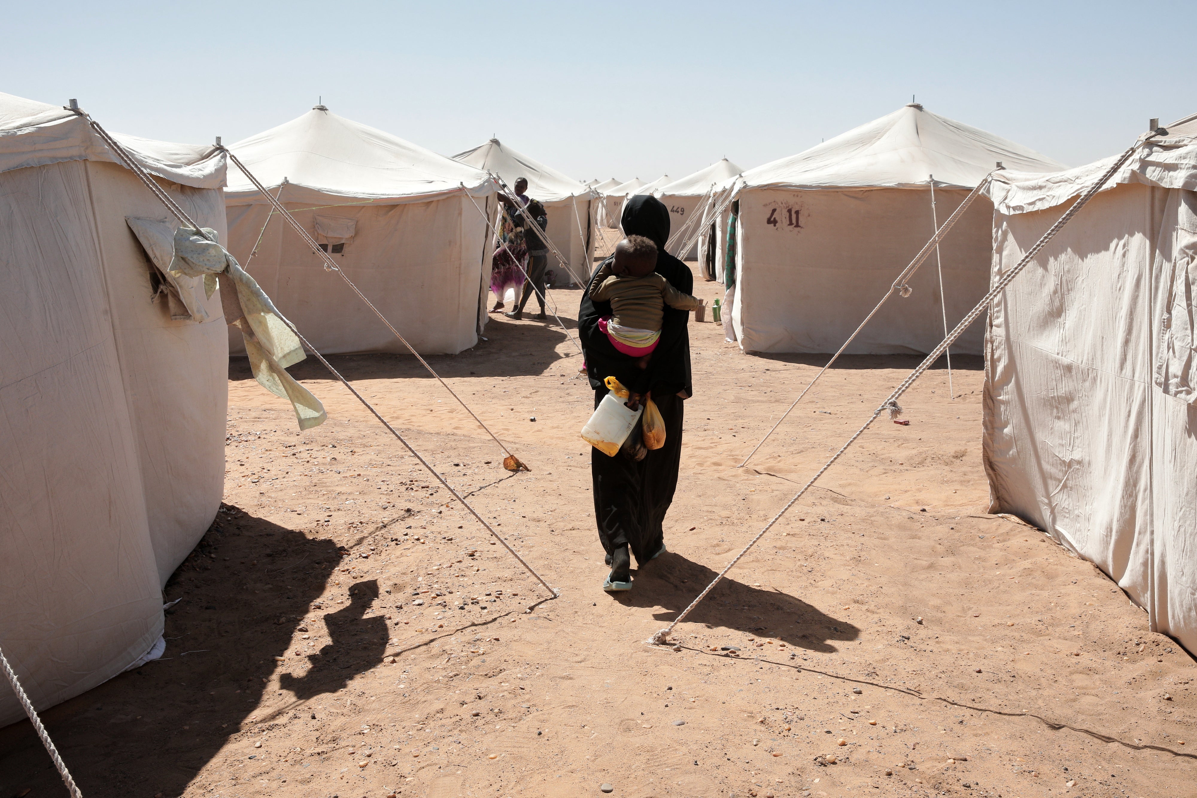 A Sudanese woman displaced from El-Fasher carries her child as she walks between tents at El-Afadh camp in Al Dabbah, in Sudan's Northern State, November 16, 2025. 