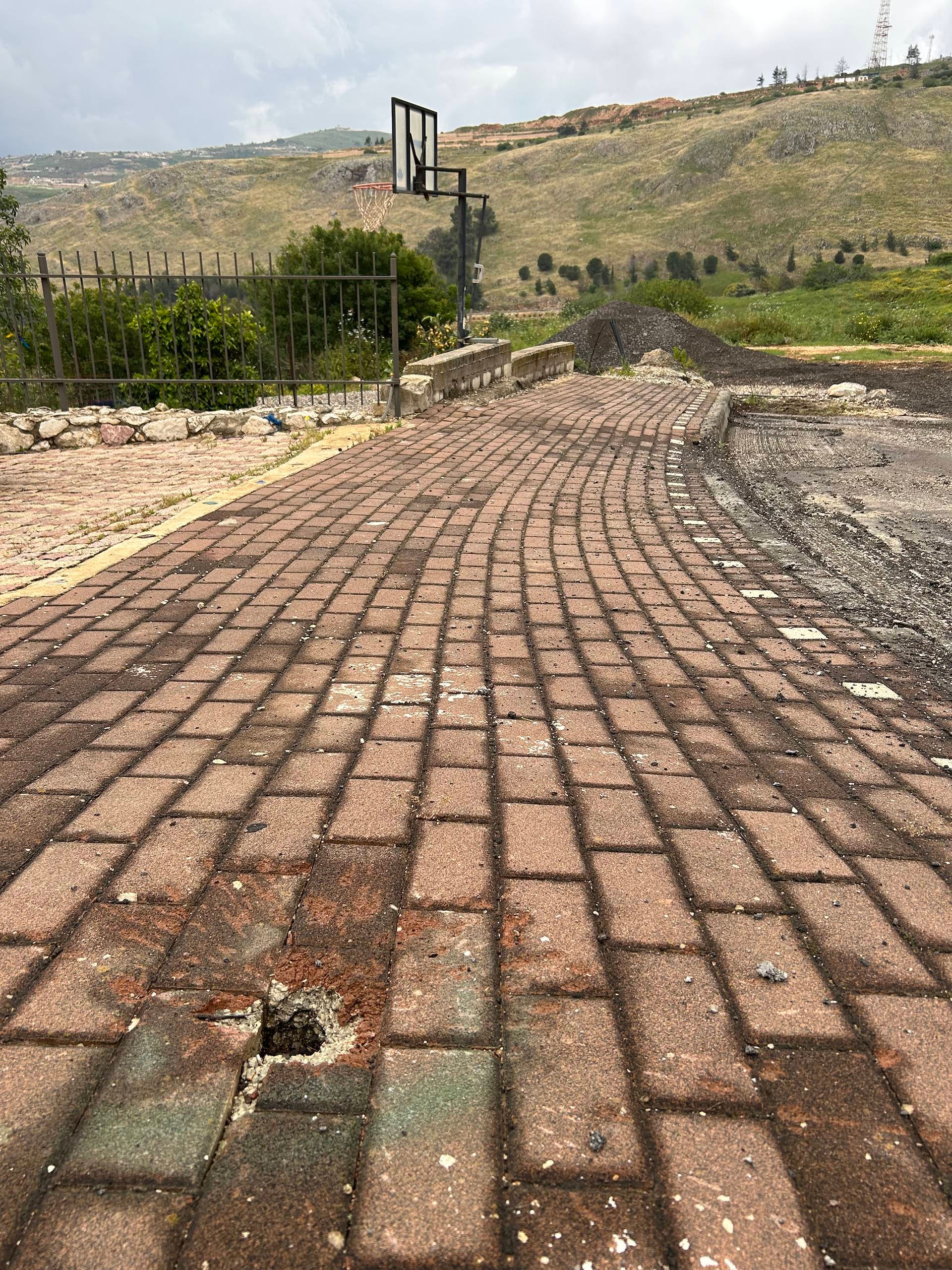 A submunition impact crater outside Hagai’s house where his son plays basketball, May 2026.