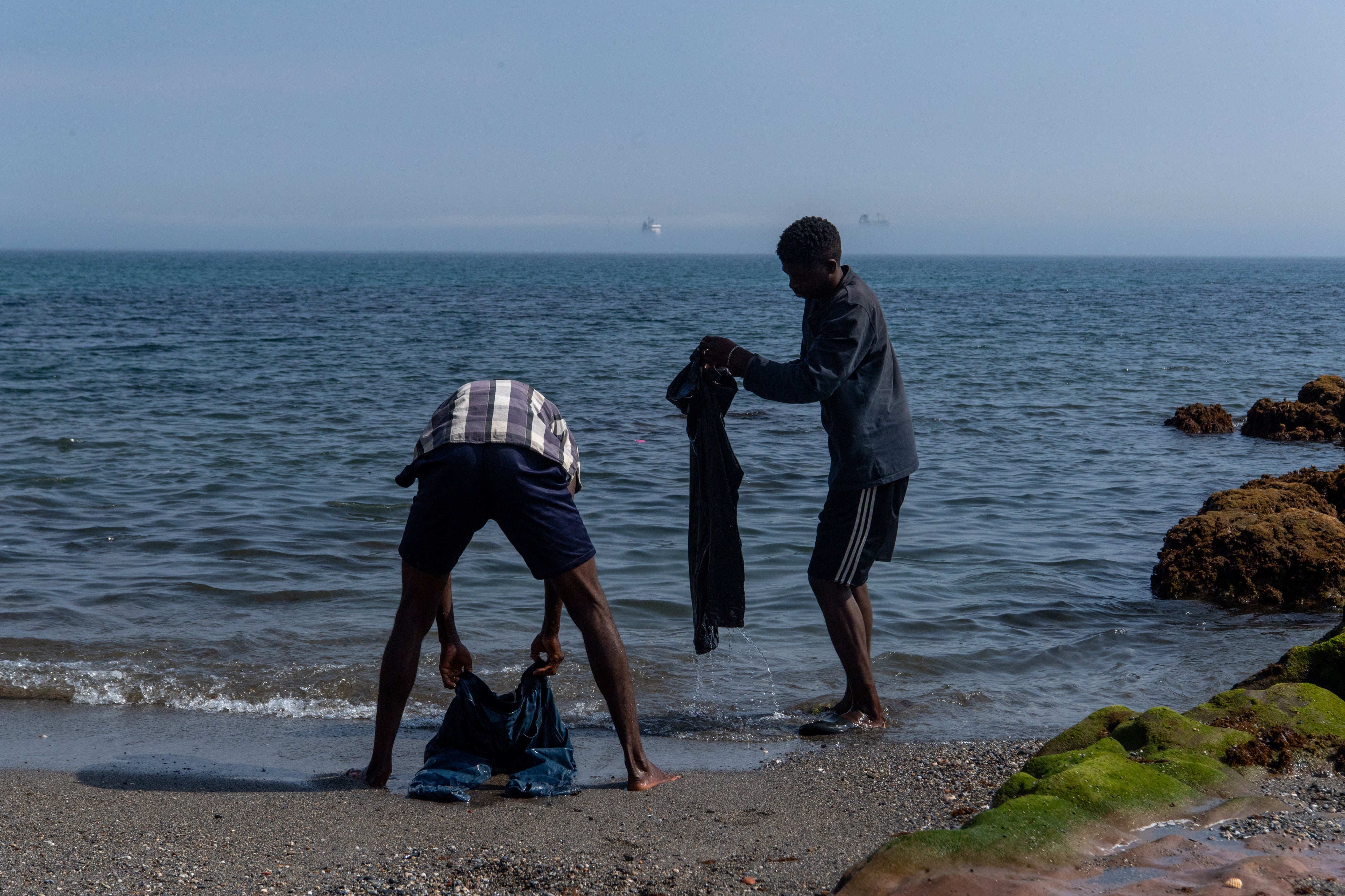 Two of the migrants and asylum seekers who arrived in Ceuta in July 2026 wash their clothes in the sea at Trampolín beach, August 2026. 