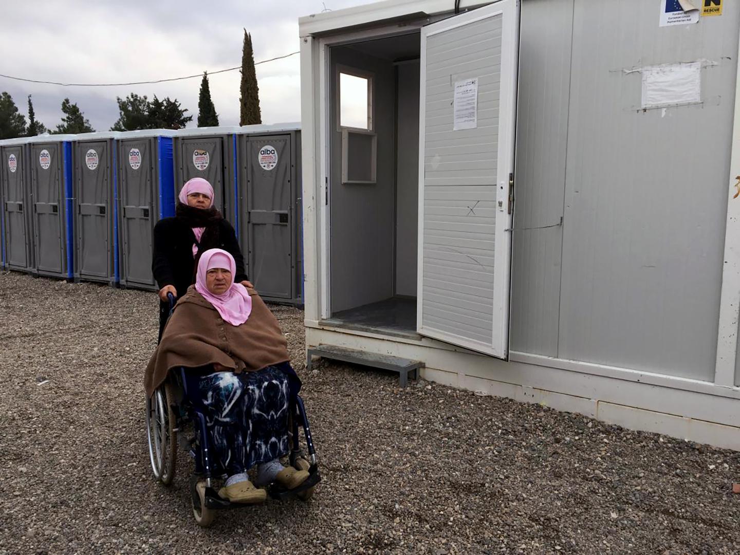 Naima, a 70-year-old woman with a disability from Aleppo, Syria, with her daughter, Hasne, in front of the shower area in Cherso camp, Thessaloniki, Greece. The showers are not accessible for people who use a wheelchair. 