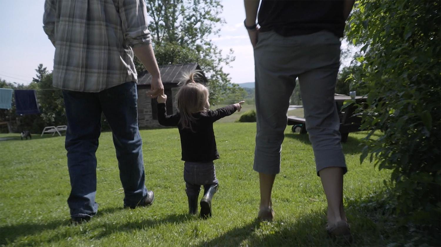 A two-and-a-half-year old born with intersex traits walks with her parents in their garden. The parents have decided to defer all medically unnecessary surgeries until their child can decide for herself.