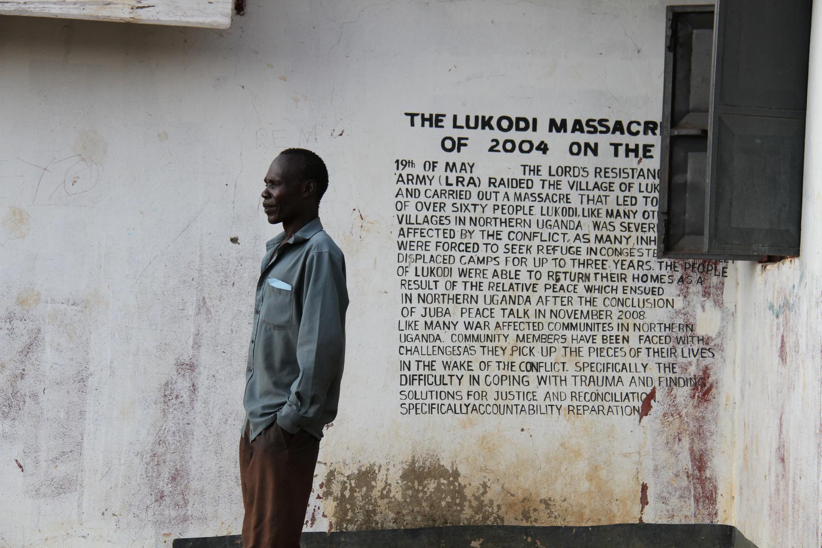 A community member in Lukodi stands next to a memorial of a May 19, 2004 massacre, one of the atrocities for which Dominic Ongwen is facing charges before the International Criminal Court. Over 4,000 victims are participating in the trial. ©2016 G. GT.