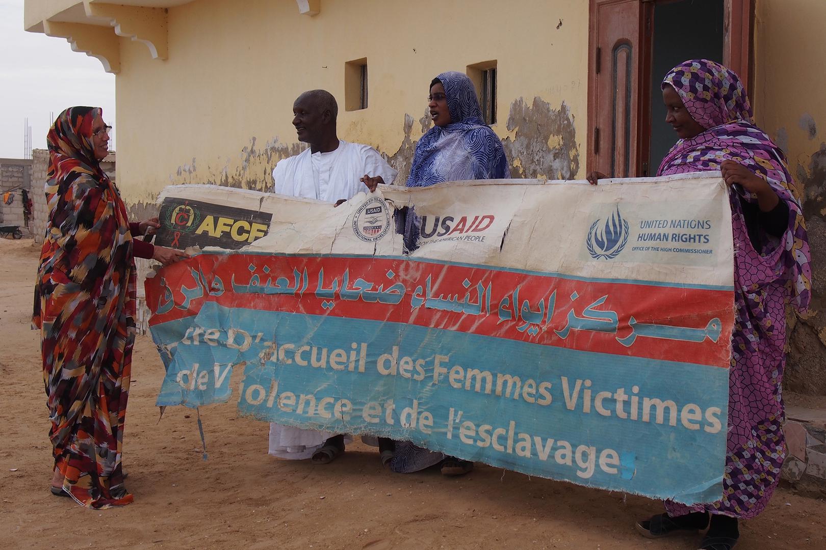 Aminetou Ely and staff of the center of the Association of Women Heads of Family in Dar Naim, with a banner for “The Reception Center for Women Victims of Violence and Slavery.” 