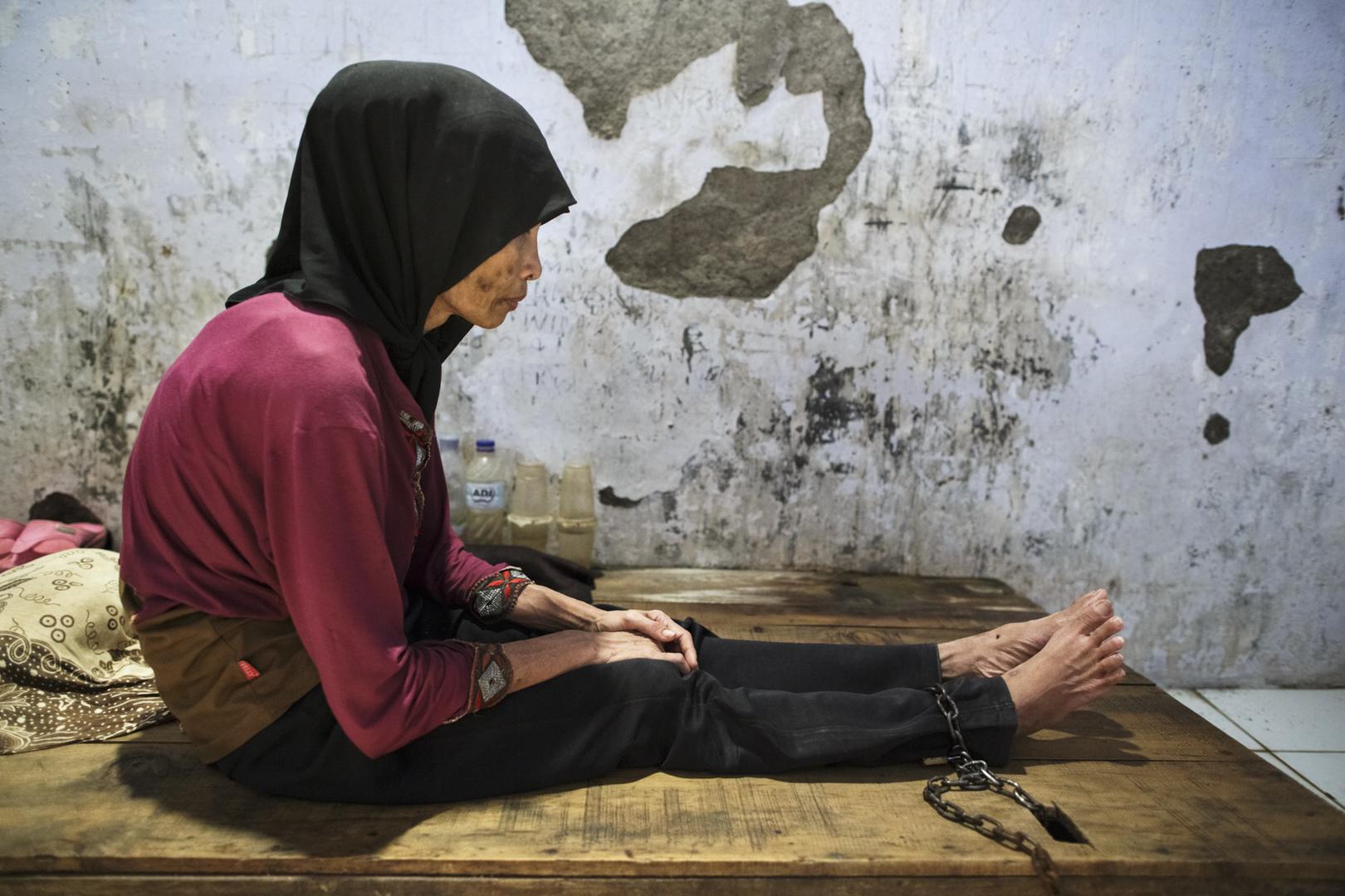 Une femme présentant un handicap psychosocial – réel ou perçu – est assise sur un lit en bois auquel l’un de ses pieds est enchaîné, dans le centre de « guérison par la foi » de Bina Lestari, à Brebes (Java central), en Indonésie.