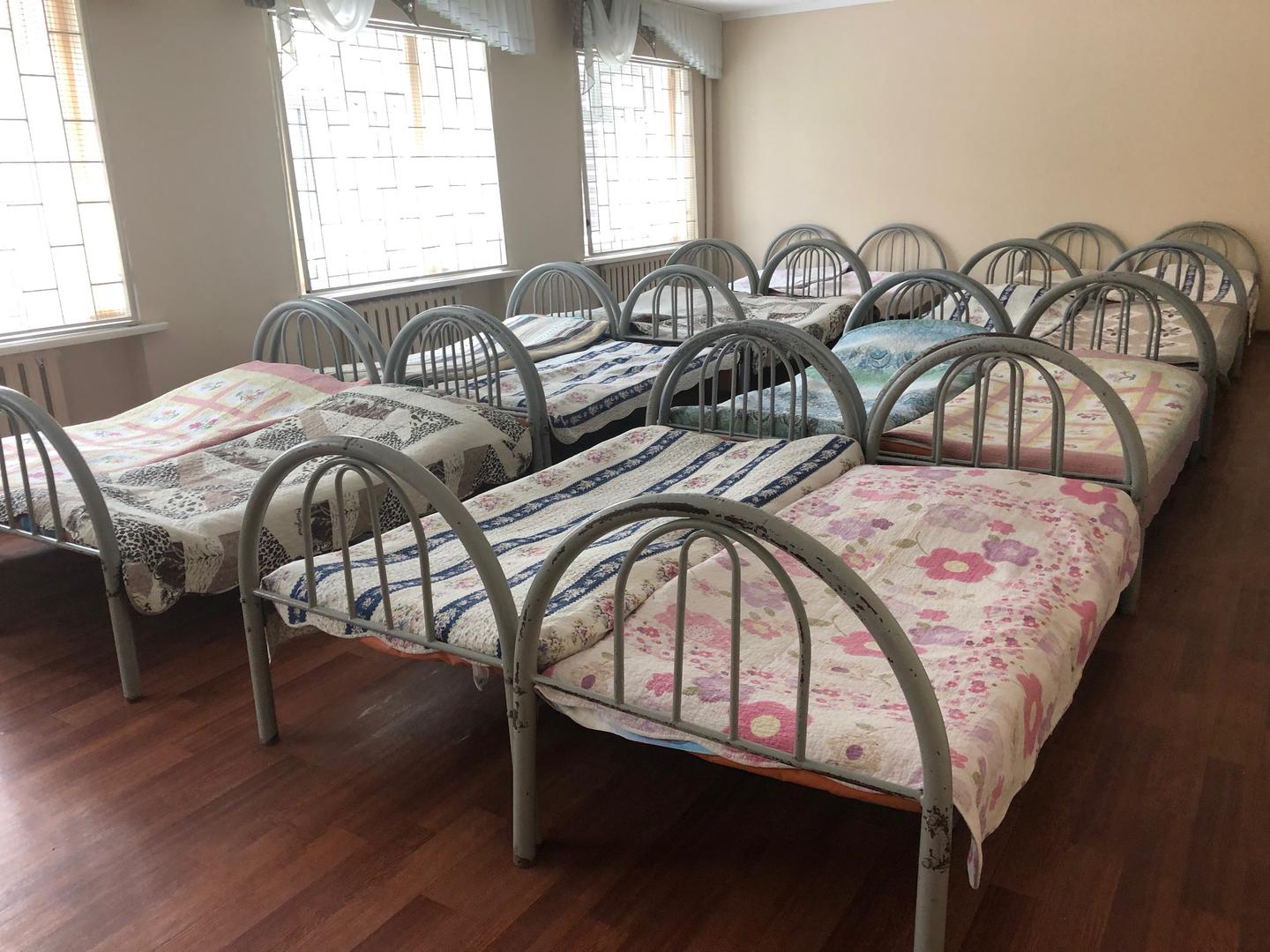 Шестнадцать кроватей, заполняющих комнату с зарешеченными окнами, в интернате для детей с инвалидностью. 