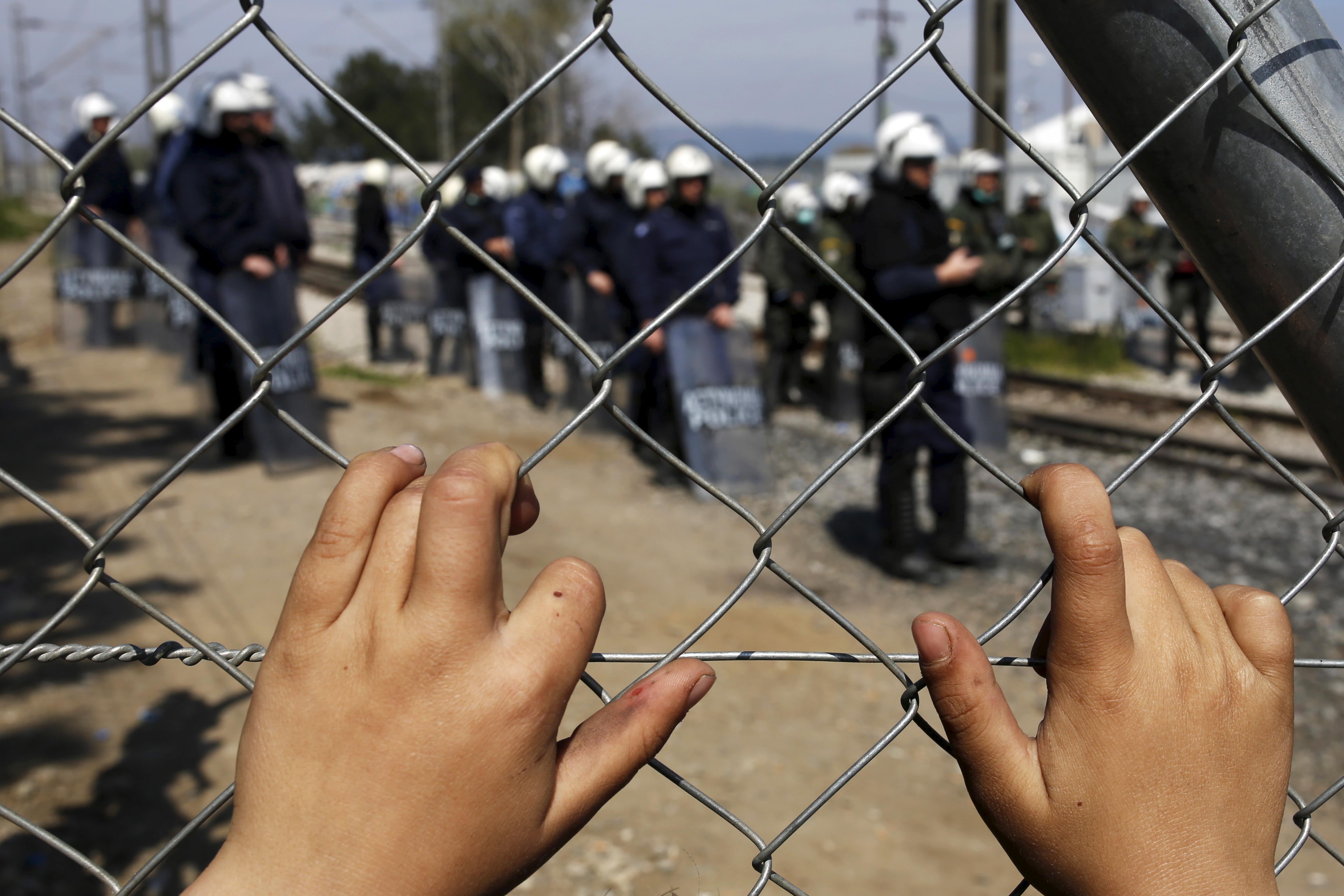 Ein Kind hält sich an einem Zaun fest, während griechische Polizisten ein Flüchtlingscamp bewachen.