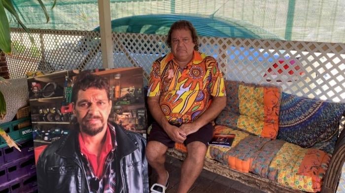 A man sits on a couch next to a large photo of his brother