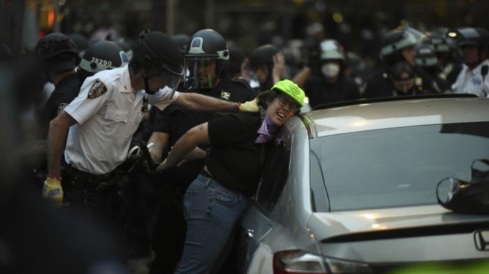 Police push a woman in a green hat against a car and handcuff her
