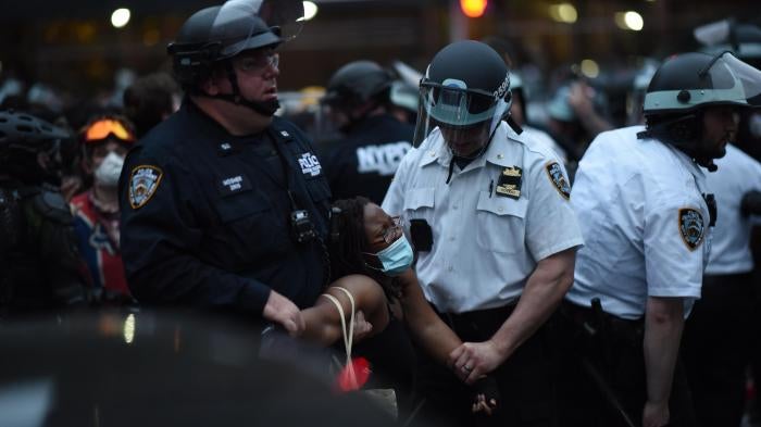 A Black woman in a mask is forcibly detained by two police officers