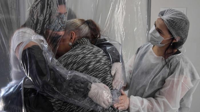 Two women in facemasks hug through a plastic curtain while a medical worker looks on