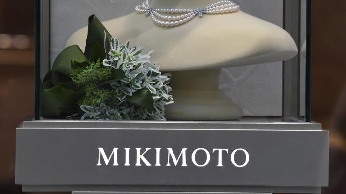 A pearl necklace is displayed over a gray sign that reads "Mikimoto"