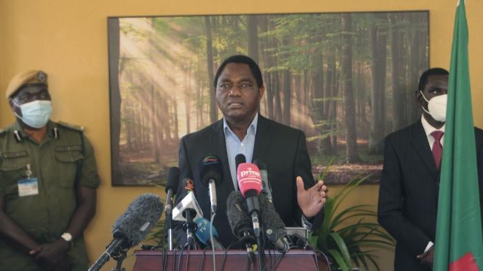 A man stands at a podium flanked by two people in face masks