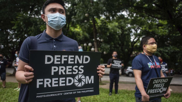 People in face masks hold signs that read "Defend Press Freedom"