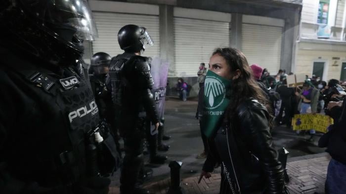 A woman wearing a green bandana over her face stands in front of a police officer with riot gear
