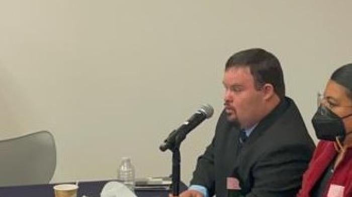 A man seated at a table speaks into a microphone