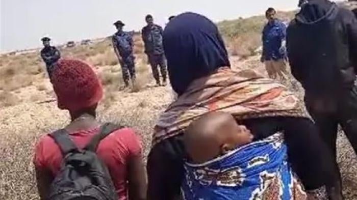 A group of Black migrants and asylum seekers of multiple African nationalities, including a woman and her baby, stranded in the desert for days after expulsion from Tunisia, stand in the buffer zone at the Tunisia-Libya border facing an Al Jazeera news crew and Libyan soldiers, July 11, 2023.