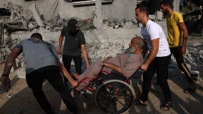 Three men help a man in a wheelchair move amid the rubble and destruction in the southern Gaza Strip on October 22, 2023. 