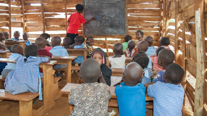 Young children sit in a rural classroom while a teacher writes on a blackboard