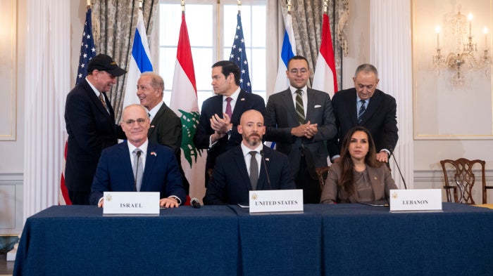 Three people sit at a table with the plaque cards for the US, Israel, and Lebanon while others behind them clap.