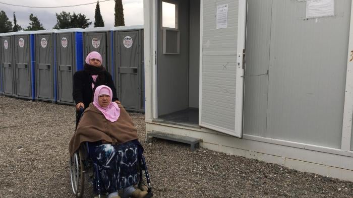 Naima, 70, an older woman with a disability from Aleppo, Syria, with her daughter Hasne, in front of the shower area in Cherso camp, Thessaloniki. The showers are not accessible for people who use a wheelchair.