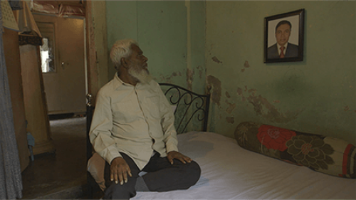 Ruhul Amin Chowdhury sits on a bed looking up at a picture of his missing son, Adnan Chowdhury, hanging on the wall.
