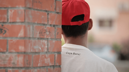 Closeup from behind of a teenager in a baseball cap and tshirt leaning against a brick column