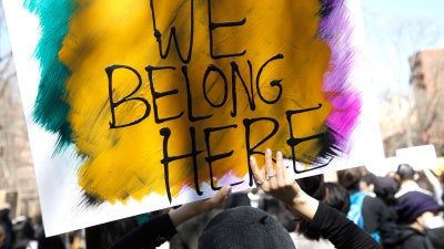 People demonstrate at a rally in Chinatown, New York City. Someone is pictured holding a painted sign reading "We Belong Here" with yellow, blue, pink, and black colors..