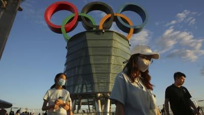 People in masks stand in front a sculpture of the Olympic rings