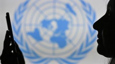 A woman looks at her cell phone in front of the United Nations logo displayed on a computer screen.