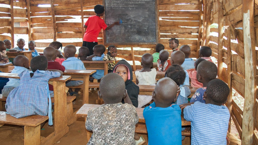 Young children sit in a rural classroom while a teacher writes on a blackboard