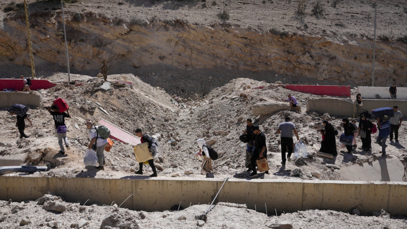 أشخاص يحملون أمتعتهم أثناء عبورهم إلى سوريا سيرا على الأقدام عبر حفرة تسببت بها ضربات جوية إسرائيلية تهدف إلى قطع طريق بيروت دمشق السريع عند معبر المصنع، شرق وادي البقاع، لبنان، السبت في 5 أكتوبر/تشرين الأول 2024.