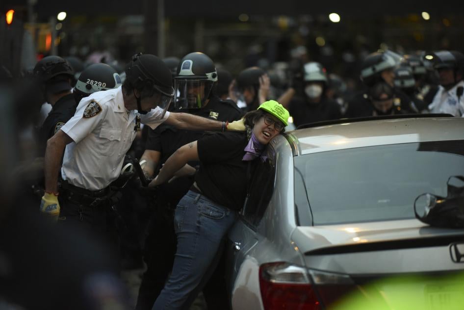 Police push a woman in a green hat against a car and handcuff her