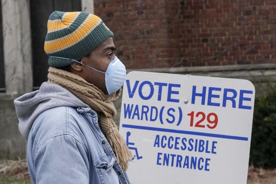 A man in a beanie and face mask stands in front of a sign that says "Vote Here"