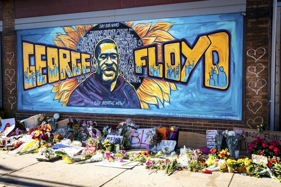 A mural dedicated to George Floyd and honoring the black lives matter movement, showing a depiction of George Floyd's face with his name and supporters of the movement against a blue painted background. Flowers and handwritten signs adorn the sidewalk beneath the mural.