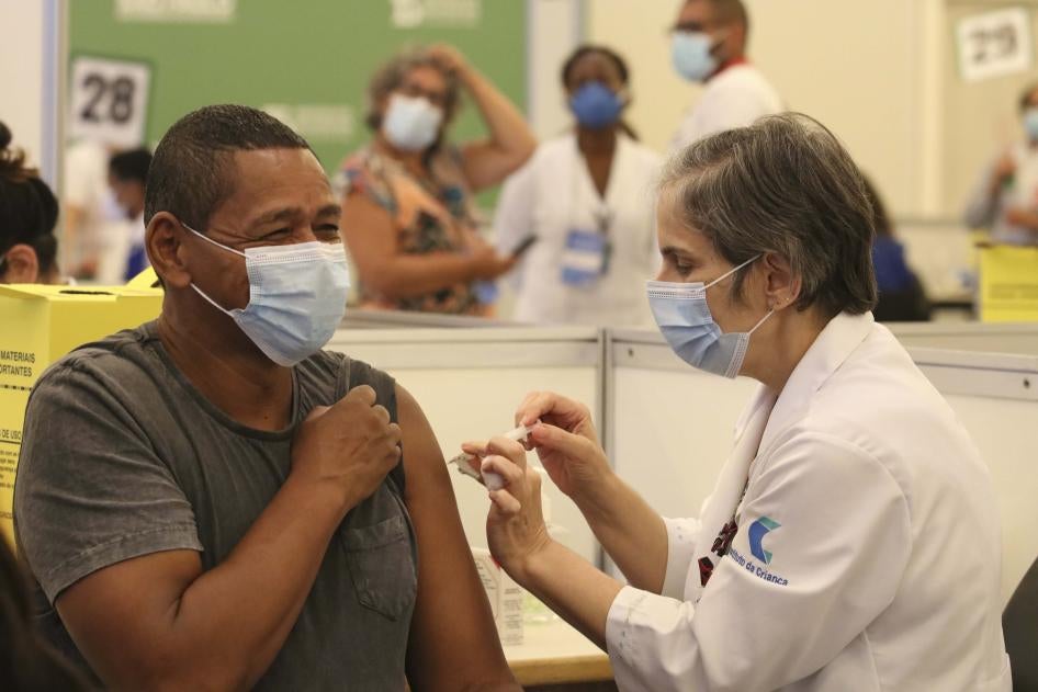 A white female healthcare professional wearing a mask, applying the covid-19 vaccine to a black male healthcare professional, wearing a mask, who is smiling