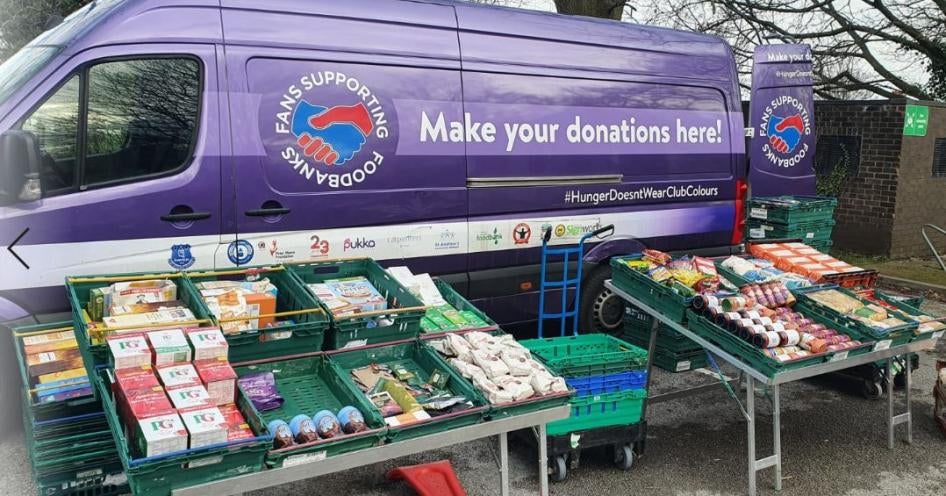 A mobile food pantry with food collected by Fans Supporting Foodbanks and other local anti-poverty groups set up at at St. Mary’s Millennium Centre, Liverpool, UK. Photo shows a purple van with cases of food set up in front of it.
